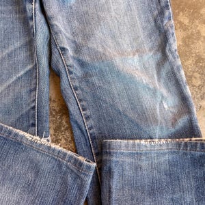 Pu&ograve; includere: Primo piano di jeans di denim blu chiaro con dettagli usurati. I jeans mostrano segni di usura, con aree sbiadite e bordi sfilacciati. Le cuciture sono visibili e il tessuto ha un aspetto strutturato.