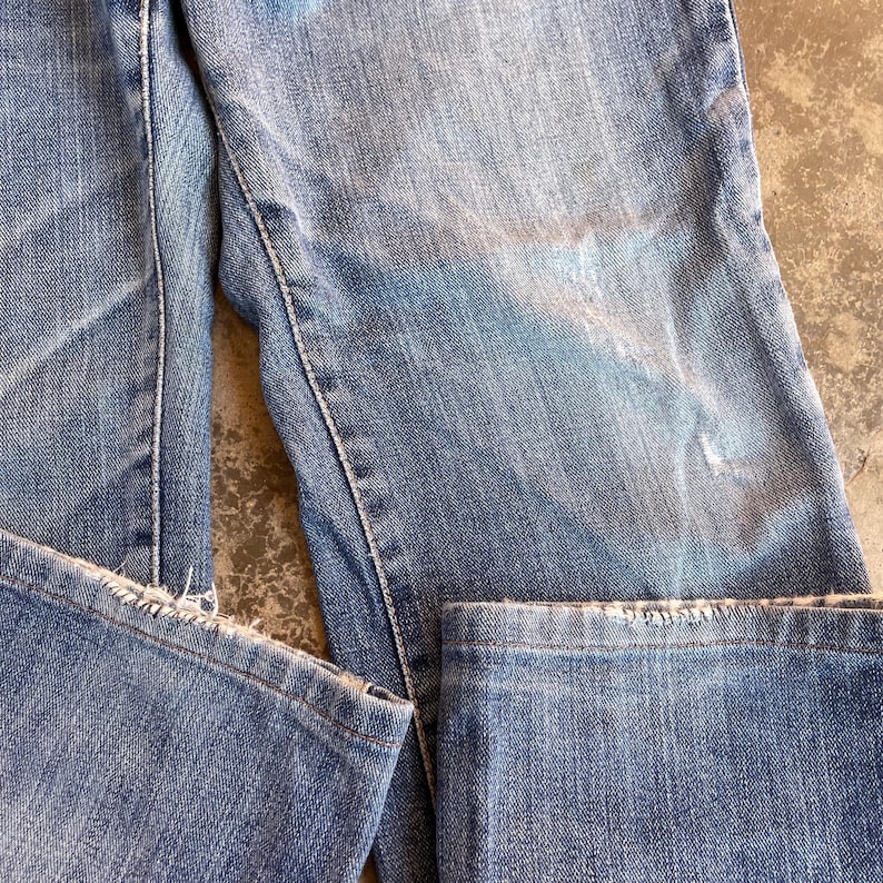 Pu&ograve; includere: Primo piano di jeans di denim blu chiaro con dettagli usurati. I jeans mostrano segni di usura, con aree sbiadite e bordi sfilacciati. Le cuciture sono visibili e il tessuto ha un aspetto strutturato.