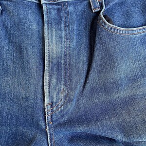 Op de afbeelding: Close-up van een blauwe spijkerbroek, met de textuur en stikdetails. De afbeelding toont de voorkant van de spijkerbroek, inclusief de knoopsluiting, de zak en de naden. De denim heeft een vervaagde uitstraling.