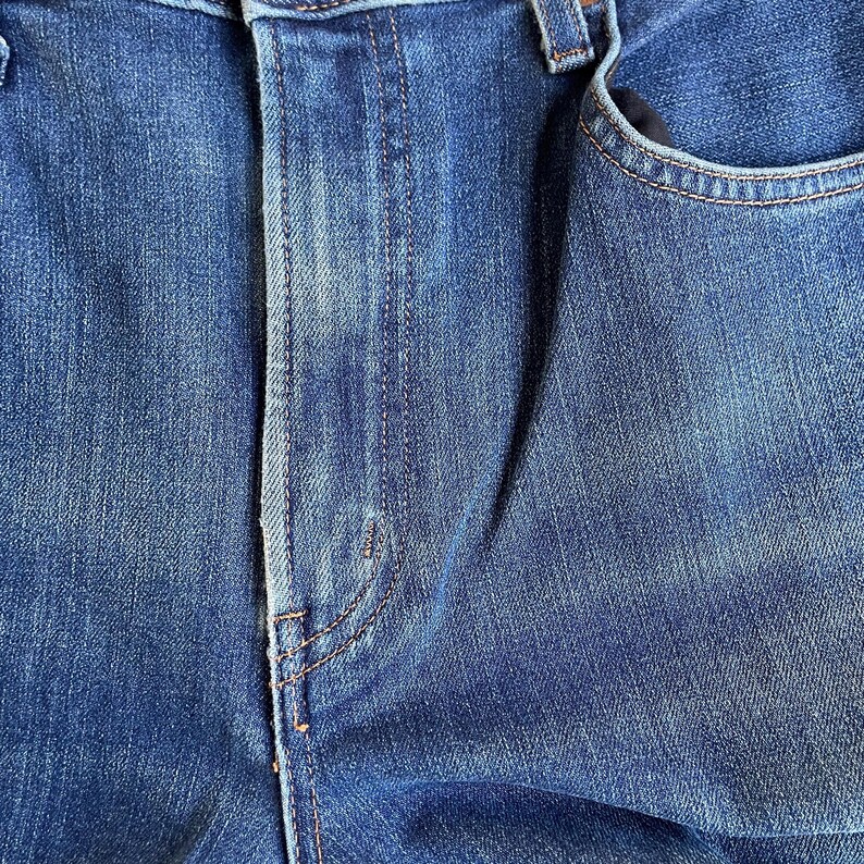Op de afbeelding: Close-up van een blauwe spijkerbroek, met de textuur en stikdetails. De afbeelding toont de voorkant van de spijkerbroek, inclusief de knoopsluiting, de zak en de naden. De denim heeft een vervaagde uitstraling.