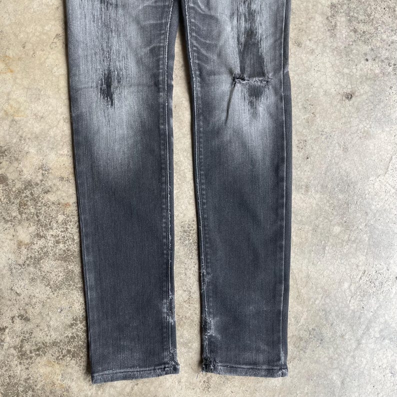 May include: Faded black denim jeans with distressed details. The jeans have a ripped knee and frayed hems, giving them a worn look. The fabric shows areas of lighter fading and wear.
