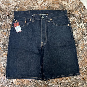 May include: Dark wash denim shorts with a button closure, five pockets, and copper-colored rivets. The shorts have a straight leg design and a small tag attached to the front. The stitching is a contrasting tan color.