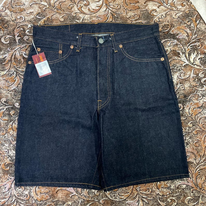 May include: Dark wash denim shorts with a button closure, five pockets, and copper-colored rivets. The shorts have a straight leg design and a small tag attached to the front. The stitching is a contrasting tan color.
