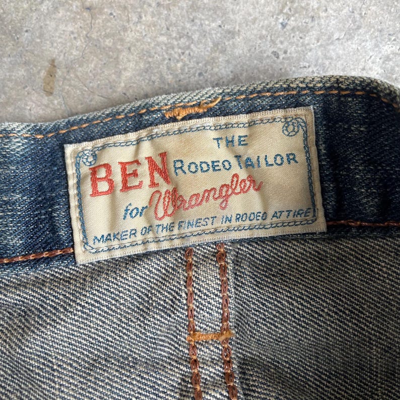 May include: Close-up of a denim garment with a rectangular tag. The tag reads "BEN for Wrangler" in red script, with additional text in a serif font. The tag is bordered with a blue and white design. The garment has visible stitching.