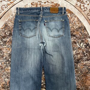 May include: A pair of blue denim jeans featuring a faded wash, two back pockets with a signature arcuate stitch design, a brown leather patch on the waistband embossed with the text EDWIN, and a small red tab on the right back pocket.
