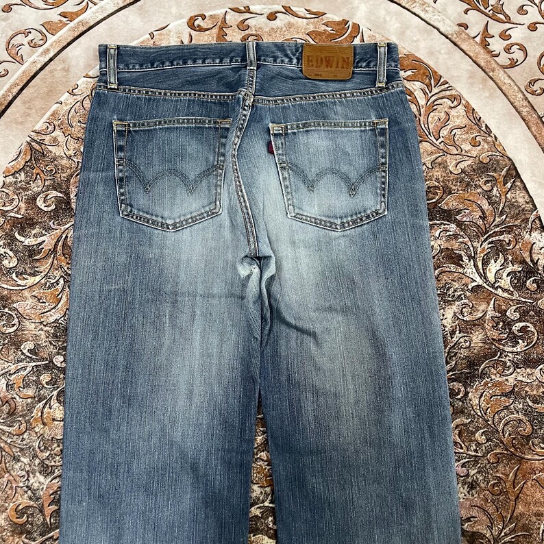 May include: A pair of blue denim jeans featuring a faded wash, two back pockets with a signature arcuate stitch design, a brown leather patch on the waistband embossed with the text EDWIN, and a small red tab on the right back pocket.