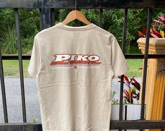 Può includere: Una maglietta beige con la scritta "PIKO" in lettere rosse e arancioni sul retro. Sotto, il testo "Hawaiian Longboard Wear" è in bianco. La maglietta è appesa a una gruccia di legno.
