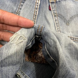 May include: Light blue denim jeans featuring a distressed, frayed area near the seam. The fabric displays a classic twill weave pattern with visible stitching details and a small red fabric tag attached to the rear pocket area of the garment.
