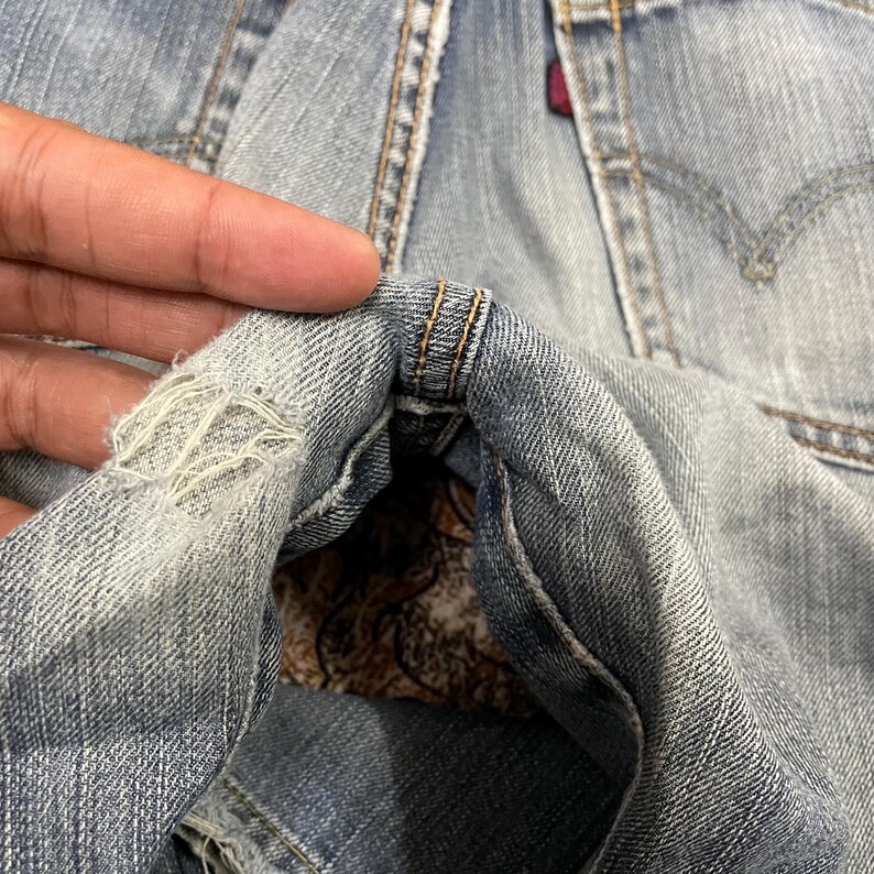 May include: Light blue denim jeans featuring a distressed, frayed area near the seam. The fabric displays a classic twill weave pattern with visible stitching details and a small red fabric tag attached to the rear pocket area of the garment.
