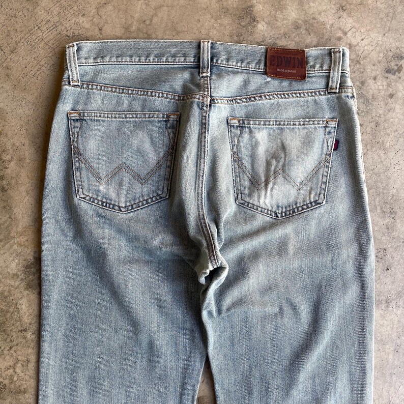 May include: Pale blue denim jeans with a classic five-pocket design. The back pockets have a distinctive W-shaped stitch. A brown leather patch with the brand name "EDWIN" is visible at the waistband. The jeans have a straight leg cut.
