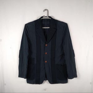 May include: A dark blue blazer with a three-button closure and two patch pockets. The blazer has a unique design with vertical panels of fabric.