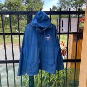 May include: A blue hooded jacket featuring the Toronto Maple Leafs logo. The jacket has a zip closure, two front pockets, and a drawstring hood. It is hanging on a black metal fence. The jacket is a vibrant shade of blue.