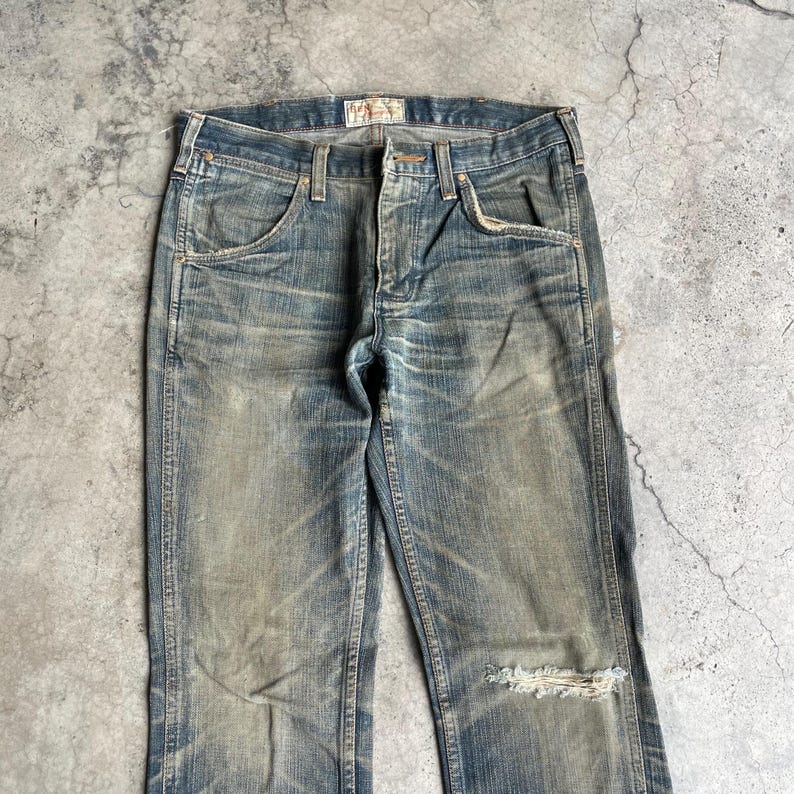 May include: Faded blue denim jeans with a distressed look. Features include classic five-pocket styling, a button and zipper closure, and a ripped knee. The jeans have a worn-in appearance.