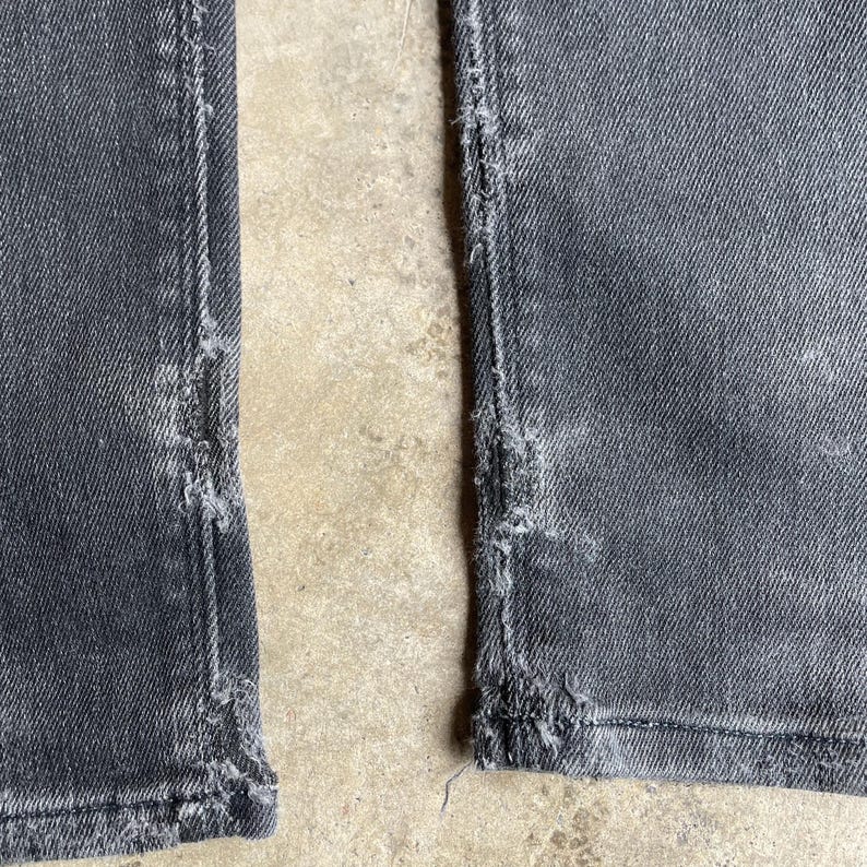 May include: Close-up of the lower portion of a pair of gray denim jeans. The jeans have distressed detailing along the seams and hems, with frayed edges and visible wear. The fabric appears faded and worn.