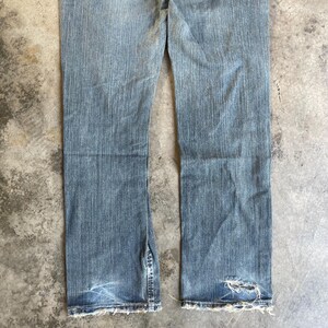 May include: Light blue denim jeans with a distressed look. The jeans have a split at the bottom of the leg and a frayed hem. There is a hole in the right leg near the hem. The jeans are worn and faded.