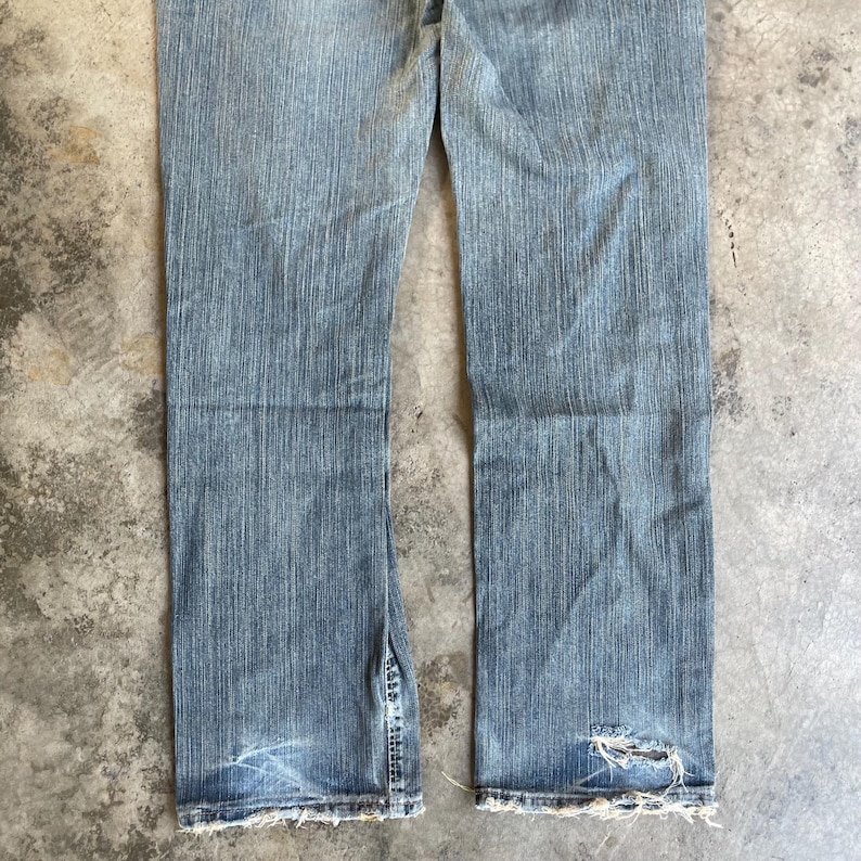 May include: Light blue denim jeans with a distressed look. The jeans have a split at the bottom of the leg and a frayed hem. There is a hole in the right leg near the hem. The jeans are worn and faded.