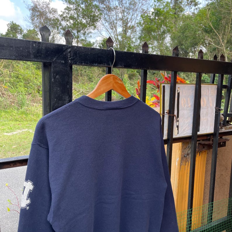 May include: A navy blue sweatshirt hangs on a wooden hanger. The sweatshirt has a white graphic on the left sleeve. The background includes a black metal fence and green foliage.