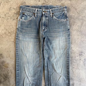 May include: Faded blue denim jeans with a classic five-pocket design. The jeans have a button closure and a straight leg fit. The denim has a worn look with lighter areas on the thighs and knees, giving them a vintage appearance.