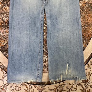 May include: Light blue denim jeans with a distressed, frayed hem and a tear near the knee. The jeans have a classic five-pocket design and a faded wash, giving them a worn-in look.