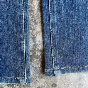 Op de afbeelding: Close-up van een blauwe spijkerbroek, met de naden en zoom. De denim heeft een vervaagde uitstraling, met zichtbare stiksels in een contrasterende kleur. De stof lijkt stevig en goed geconstrueerd, wat duurzaamheid suggereert.