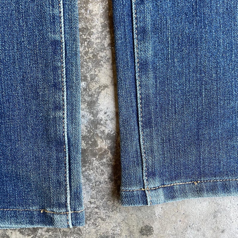 Op de afbeelding: Close-up van een blauwe spijkerbroek, met de naden en zoom. De denim heeft een vervaagde uitstraling, met zichtbare stiksels in een contrasterende kleur. De stof lijkt stevig en goed geconstrueerd, wat duurzaamheid suggereert.