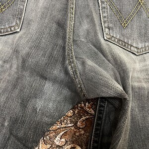 May include: Close-up of gray denim jeans with yellow stitching details. The back pockets have a classic design. The jeans are slightly faded, showing signs of wear. The fabric has a textured appearance.