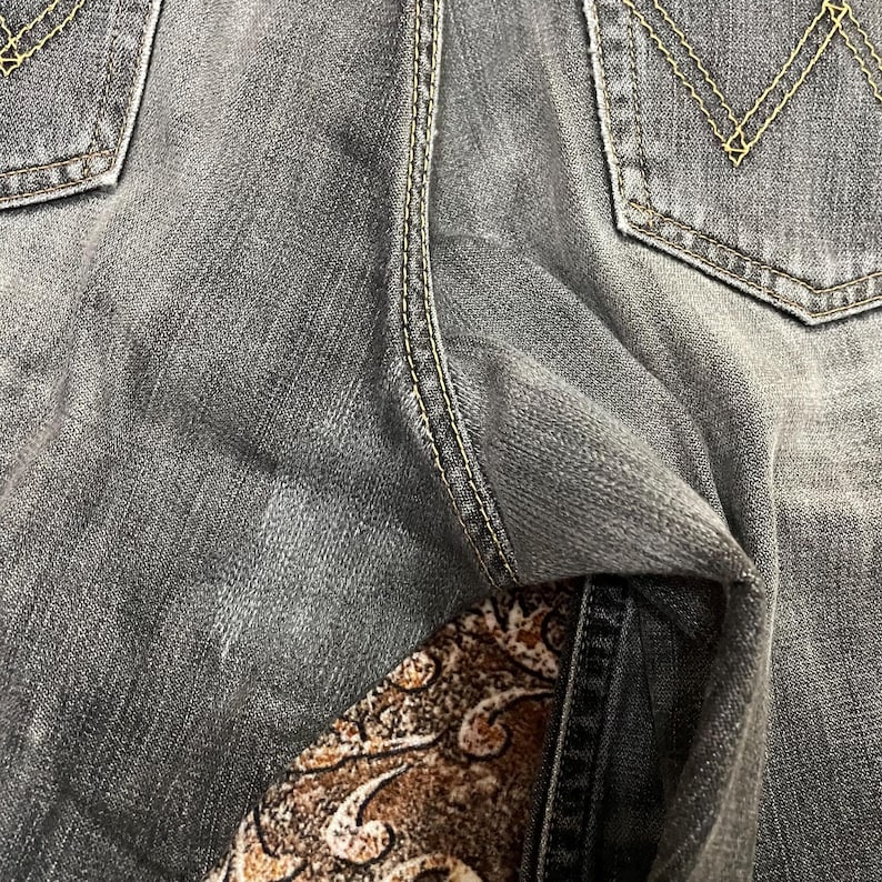 May include: Close-up of gray denim jeans with yellow stitching details. The back pockets have a classic design. The jeans are slightly faded, showing signs of wear. The fabric has a textured appearance.