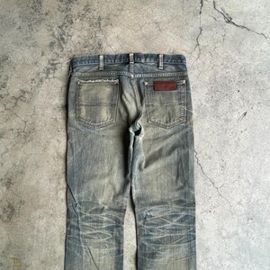 May include: Faded blue denim jeans with a distressed look. The jeans feature two back pockets, a brown leather patch, and a classic five-pocket design. The denim has a worn appearance with visible wear marks and fading.
