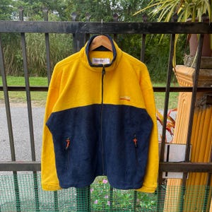 Op de afbeelding: Een geel en blauw fleecejack met een ritssluiting. Het jack heeft het woord "Nautica" en "Competition" geborduurd op de voorkant.