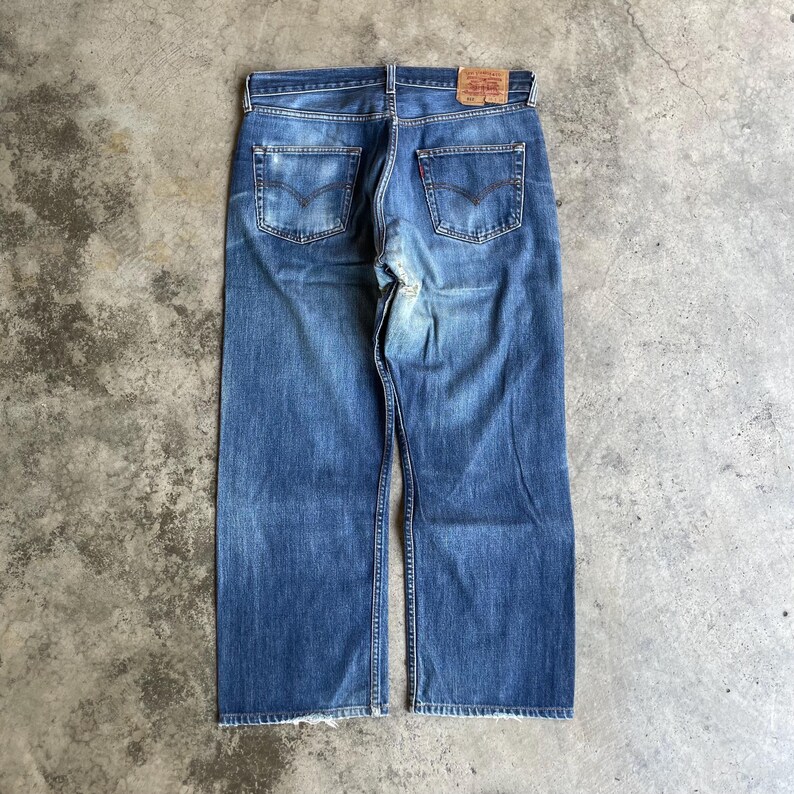 May include: Faded blue denim jeans with a classic five-pocket design and a leather patch. The jeans have a straight leg cut and are laid flat, displaying the back pockets and the brand's red tab. The jeans are approximately 100 cm in length.