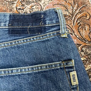 May include: A pair of blue denim jeans featuring a dark wash, yellow contrast stitching, a belt loop, and a small rectangular beige fabric tag on the back pocket with the word EDWIN printed in black capital letters on the fabric label.