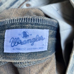 May include: Close-up of a denim jacket collar with a light blue Wrangler label. The label reads "Wrangler AUTHENTIC WESTERN WEAR" and features a silhouette. The denim is faded with visible stitching.
