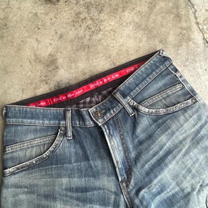 K&ouml;nnte beinhalten: Blaue Jeans aus Denim mit klassischem F&uuml;nf-Taschen-Design. Die Jeans haben einen Knopf- und Rei&szlig;verschluss, mit einem roten Bund mit dem Text "EDGE or Blue Jeans". Die Jeans sind verblichen.
