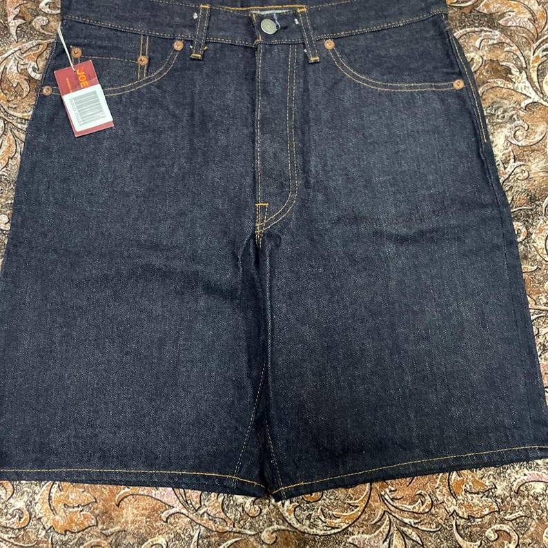 May include: Dark wash denim shorts with classic five-pocket styling. The shorts feature a button and zipper closure, belt loops, and contrast stitching. A small tag is attached to the shorts. The shorts are a medium length.