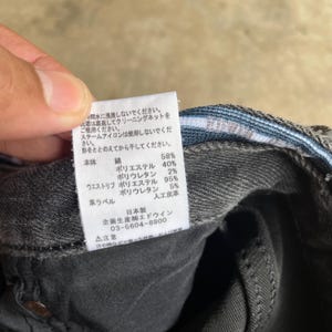 May include: Close-up of a tag attached to a pair of dark gray denim jeans. The tag has Japanese text and details the fabric composition: 58% cotton, 40% polyester, and 2% polyurethane.