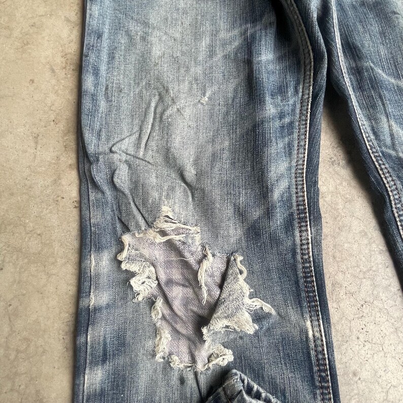 K&ouml;nnte beinhalten: Nahaufnahme einer Jeans aus blauem Denim mit Used-Look. Die Jeans hat ein gro&szlig;es, ausgefranstes Loch am Knie, das einen helleren Stoff darunter freilegt. Der Denim weist Gebrauchsspuren auf, mit verblassten Bereichen und subtilen Falten.