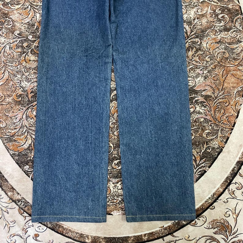 Puede incluir: Un par de vaqueros azules de mezclilla con un dise&ntilde;o de pierna ancha. Los vaqueros est&aacute;n hechos de tela de mezclilla de lavado medio y tienen un dise&ntilde;o cl&aacute;sico de cinco bolsillos. Los vaqueros est&aacute;n colocados planos sobre una superficie estampada.