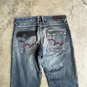 K&ouml;nnte beinhalten: Verblasste blaue Jeans mit einzigartigen Taschen-Designs. Die Ges&auml;&szlig;taschen haben eine markante rote wellenf&ouml;rmige Naht. Die Jeans haben einen Knopfverschluss und eine dunkelgraue Tasche auf der linken Seite. Die Jeans sind getragen.