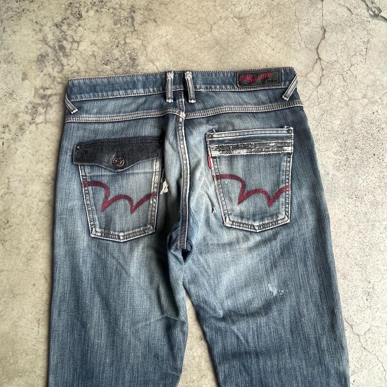 K&ouml;nnte beinhalten: Verblasste blaue Jeans mit einzigartigen Taschen-Designs. Die Ges&auml;&szlig;taschen haben eine markante rote wellenf&ouml;rmige Naht. Die Jeans haben einen Knopfverschluss und eine dunkelgraue Tasche auf der linken Seite. Die Jeans sind getragen.