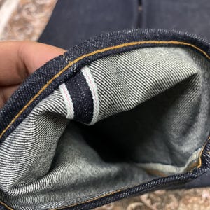 May include: Close-up of dark blue denim jeans cuff, showing the interior fabric and stitching. The cuff is folded over, revealing a white and gray striped pattern with a pink accent. The outer edge has a yellow-orange stitch.
