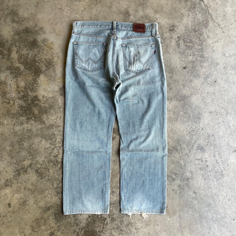 May include: Pale blue denim jeans with a classic five-pocket design. The jeans have a faded wash, a brown leather patch, and a straight leg cut. The back pockets feature a distinctive W-shaped stitching.