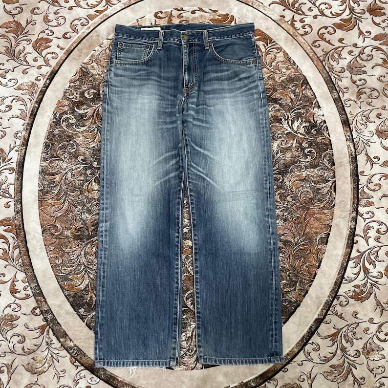 May include: A pair of blue denim jeans featuring a faded wash, a classic five-pocket design, a button fly closure, and visible stitching along the seams. The fabric displays a light blue centre with darker blue edges and a straight leg cut.