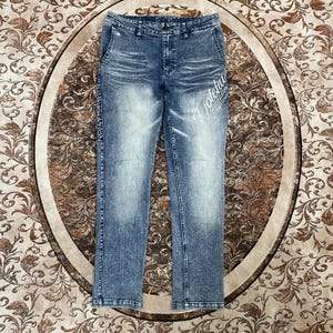 May include: Blue acid-wash denim jeans featuring a button fly, belt loops, and five pockets. The front displays white script text reading "Gotcha" and "California" on the left thigh, with "LIBERATION" printed vertically along the right side seam.