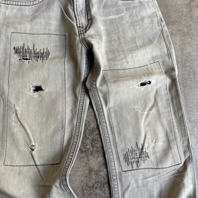 K&ouml;nnte beinhalten: Hellgraue Jeans mit Used-Details. Die Jeans hat schwarze N&auml;hte und Patches mit grafischen Designs. Es gibt auch L&ouml;cher im Stoff, die dem Used-Look entsprechen. Die Jeans hat ein klassisches F&uuml;nf-Taschen-Design.