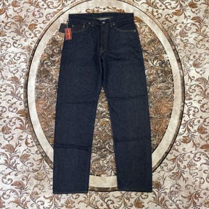 May include: Dark wash denim jeans with a classic five-pocket design. The jeans have a button fly and a small tag attached. The jeans are displayed on a patterned surface.