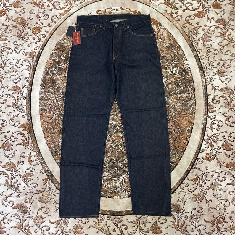 May include: Dark wash denim jeans with a classic five-pocket design. The jeans have a button fly and a small tag attached. The jeans are displayed on a patterned surface.