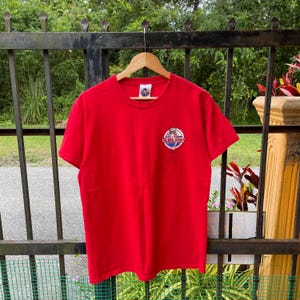 Könnte beinhalten: Ein leuchtend rotes T-Shirt mit einem kleinen kreisförmigen Logo auf der linken Brust. Das Logo zeigt den Text "Bubba Gump" in einem kreisförmigen Design. Das T-Shirt hängt an einem Holzbügel vor einem Hintergrund aus Grün.