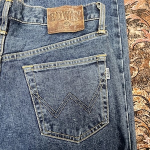 Puede incluir: Primer plano de unos vaqueros azules con un bolsillo trasero con un dise&ntilde;o de costura en forma de W. Los vaqueros tienen un parche de cuero marr&oacute;n con la marca "EDWIN" y el n&uacute;mero "403".