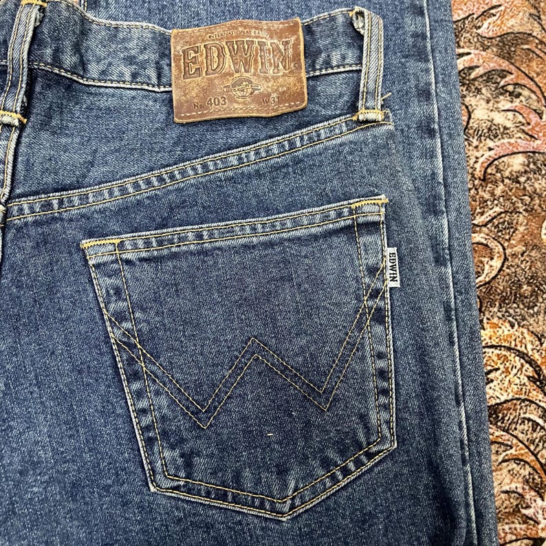 Puede incluir: Primer plano de unos vaqueros azules con un bolsillo trasero con un dise&ntilde;o de costura en forma de W. Los vaqueros tienen un parche de cuero marr&oacute;n con la marca "EDWIN" y el n&uacute;mero "403".