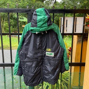 Könnte beinhalten: Schwarz-grüne Kapuzenjacke mit Reißverschluss. Die Jacke hat grüne Ärmel und Kapuze, mit einem schwarzen Körper. Auf der Vorderseite befindet sich ein kleiner Aufnäher mit dem Text "F-1 RACING". Die Jacke hängt an einem schwarzen Metallzaun.